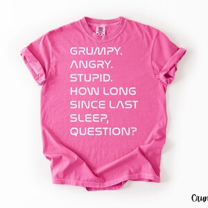 May include: A bright pink t-shirt with white text. The text reads: "GRUMPY. ANGRY. STUPID. HOW LONG SINCE LAST SLEEP, QUESTION?" The shirt has rolled-up sleeves and a crew neck. The word "Crunchberry" is in the bottom right corner.