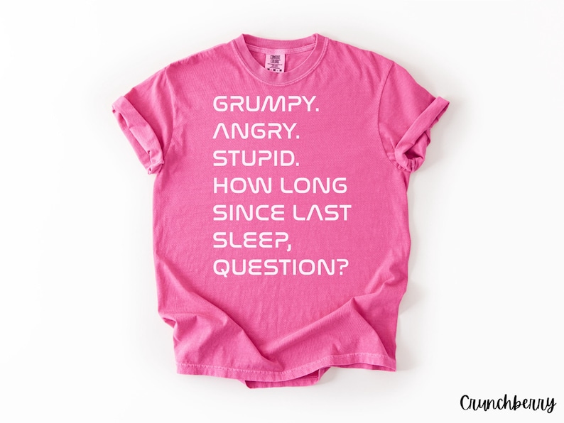 May include: A bright pink t-shirt with white text. The text reads: "GRUMPY. ANGRY. STUPID. HOW LONG SINCE LAST SLEEP, QUESTION?" The shirt has rolled-up sleeves and a crew neck. The word "Crunchberry" is in the bottom right corner.