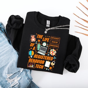 Op de afbeelding: Zwart sweatshirt met de tekst "THE LIFE OF A REGISTERED BEHAVIOR TECH" en afbeeldingen. Het ontwerp bevat een teddybeer, laptop, hersenen en andere iconen. Een zilveren ketting ligt over het sweatshirt. Het sweatshirt ligt naast een blauwe spijkerbroek.