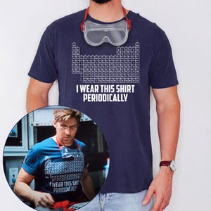 Puede incluir: Una camiseta de manga corta azul marino con un gráfico blanco de la tabla periódica de los elementos y el texto "I WEAR THIS SHIRT PERIODICALLY" impreso en letras mayúsculas blancas. La camiseta se lleva con gafas de seguridad.
