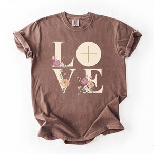 May include: Espresso-coloured t-shirt featuring the word "LOVE" in a cream font. The letters "L" and "E" are decorated with floral designs in pink, peach, and green. A cross is in the "O". The shirt is folded.
