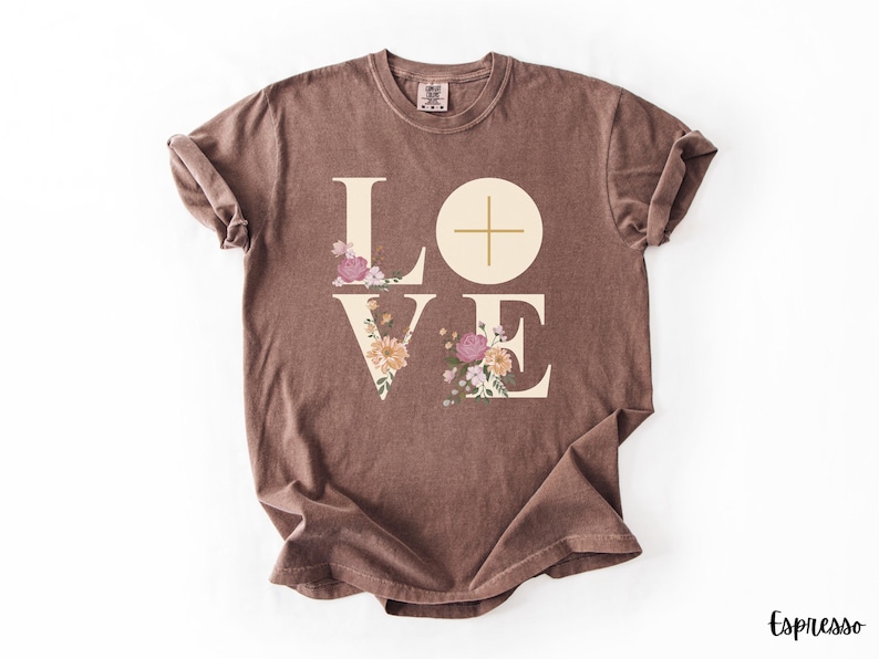May include: Espresso-coloured t-shirt featuring the word "LOVE" in a cream font. The letters "L" and "E" are decorated with floral designs in pink, peach, and green. A cross is in the "O". The shirt is folded.
