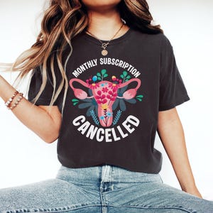 May include: Dark grey t-shirt with the text "MONTHLY SUBSCRIPTION CANCELLED" in white. The shirt features a floral uterus design in pink, green, and blue. The shirt is made of a soft material.