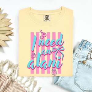 May include: A pale yellow t-shirt featuring a graphic design with vertical pink stripes, two cherries, two strawberries, and a blue bow. The text reads I need an alani in blue script font with a pink outline. The shirt has a Comfort Colors brand label.