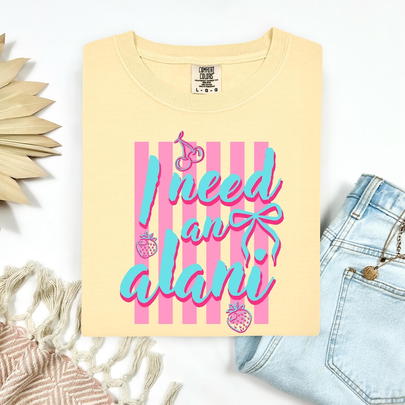 May include: A pale yellow t-shirt featuring a graphic design with vertical pink stripes, two cherries, two strawberries, and a blue bow. The text reads I need an alani in blue script font with a pink outline. The shirt has a Comfort Colors brand label.