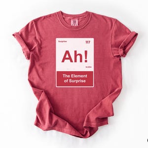 May include: A crimson short-sleeve t-shirt featuring a white rectangular graphic designed like a periodic table element. The graphic includes the text Surprise, 117, Ah!, 14.094, and The Element of Surprise in red and white lettering.
