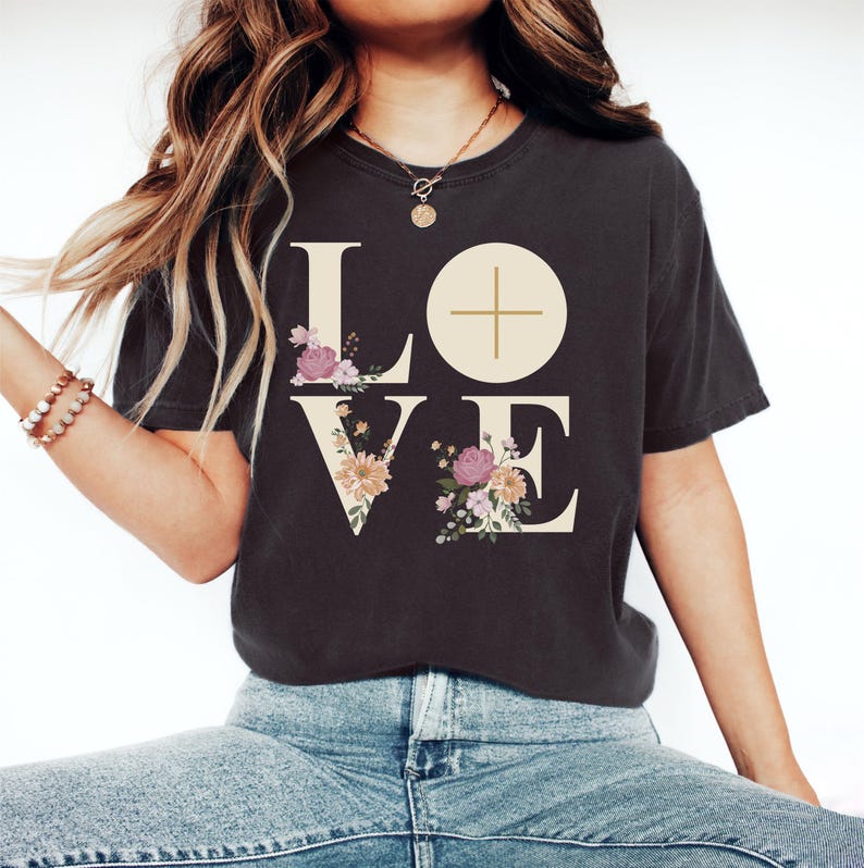 May include: Dark gray t-shirt with the word "LOVE" in large, cream-colored letters. The "O" is a circle with a cross inside. Floral arrangements in pink, purple, and yellow decorate the letters. The shirt is worn by a person wearing a gold necklace and bracelet.