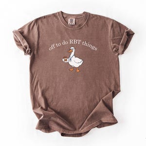 May include: A brown short-sleeve t-shirt featuring a graphic of a white goose wearing glasses and a blue lanyard while holding a coffee cup. The text above the goose reads off to do RBT things in white lettering. The shirt has a crew neckline.