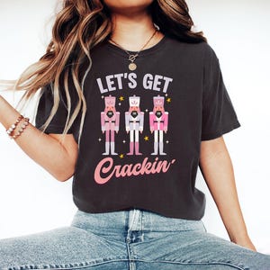 May include: Dark grey t-shirt with the text "LET'S GET Crackin'" and three nutcracker figures in pink, white, and grey. The design includes star accents. The shirt is a casual style.