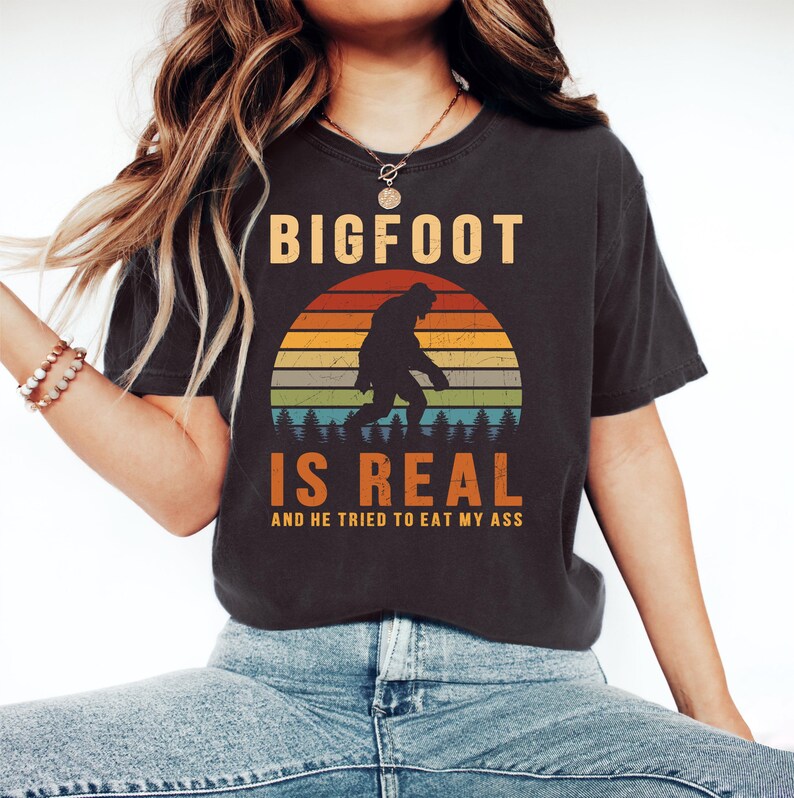 Op de afbeelding: Donkergrijs t-shirt met de tekst "BIGFOOT IS REAL" in oranje en wit. Het ontwerp toont een Bigfoot-silhouet voor een zonsondergang met oranje, gele, groene en blauwe strepen. De tekst "AND HE TRIED TO EAT MY ASS" staat eronder.
