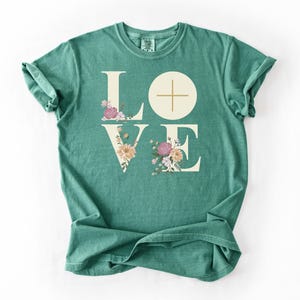 May include: A light green t-shirt featuring the word "LOVE" in a cream font. The "O" is a circle with a cross, and the letters "L" and "V" are decorated with flowers. The words "Light green" are in the bottom right corner.