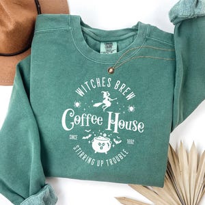 May include: A sage green crewneck sweatshirt featuring a white graphic print that reads Witches Brew Coffee House Since 1692 Stirring Up Trouble. The design includes a witch on a broomstick, a cauldron, bats, spiders, and stars.
