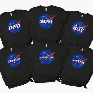 Op de afbeelding: Zwarte t-shirts met een op NASA geïnspireerd logo. Het ontwerp bevat een blauwe cirkel met witte sterren en een rode swoosh. De shirts tonen de woorden "DAD", "MOM", "BIRTHDAY BOY", "BROTHER", "SISTER" en "CUSTOM" in het wit.