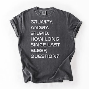 May include: Dark gray t-shirt with the text "GRUMPY. ANGRY. STUPID. HOW LONG SINCE LAST SLEEP, QUESTION?" in white block letters. The shirt has short sleeves and a crew neck.