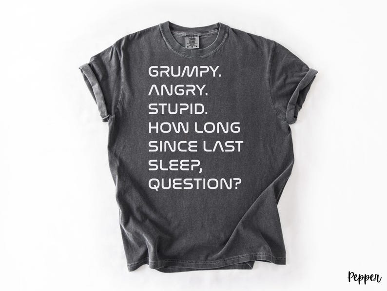 May include: Dark gray t-shirt with the text "GRUMPY. ANGRY. STUPID. HOW LONG SINCE LAST SLEEP, QUESTION?" in white block letters. The shirt has short sleeves and a crew neck.