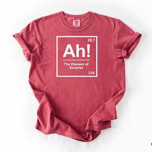 May include: Crimson t-shirt featuring the text "Ah! The Element of Surprise" in a white box. The shirt has rolled-up sleeves and a crew neck. The text includes the numbers 29,7 and 230.