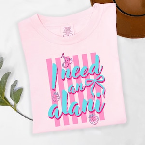 May include: A light pink short-sleeved t-shirt featuring a graphic print of vertical pink stripes, a blue bow, cherries, and strawberries. The text reads I need an alani in blue script. The shirt has a Comfort Colors brand label at the neck.