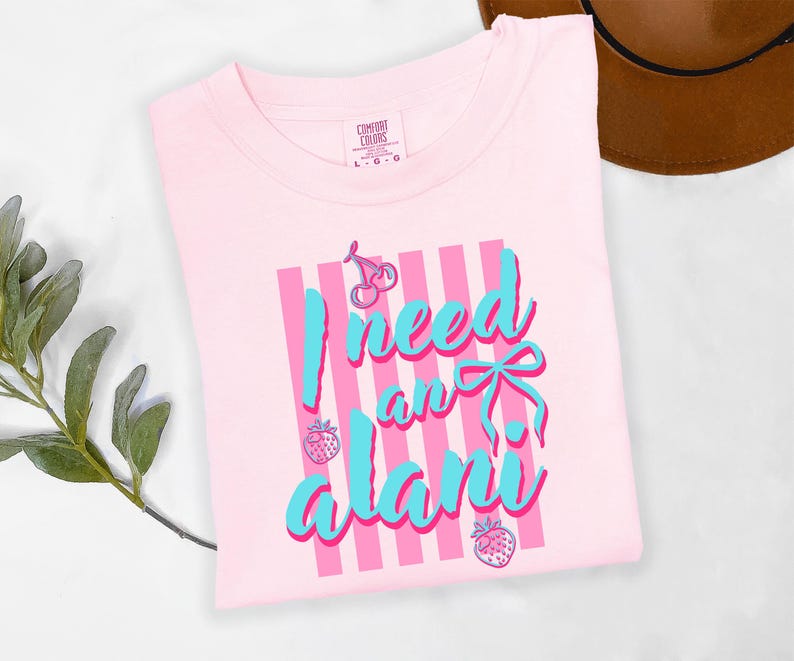 May include: A light pink short-sleeved t-shirt featuring a graphic print of vertical pink stripes, a blue bow, cherries, and strawberries. The text reads I need an alani in blue script. The shirt has a Comfort Colors brand label at the neck.