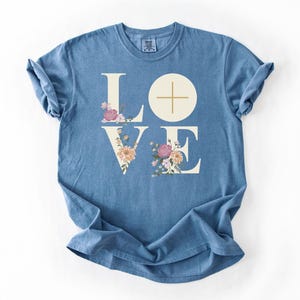 May include: A blue jean t-shirt featuring the word "LOVE" in a white serif font. The letters "L" and "E" are decorated with floral arrangements in pink, peach, and yellow. A cross is in the "O". The text "Blue Jean" is in the bottom right corner.