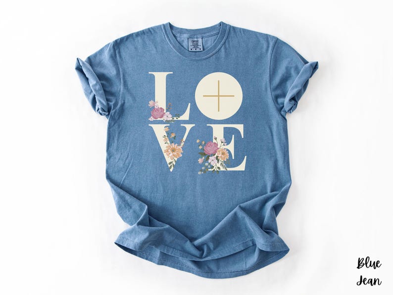 May include: A blue jean t-shirt featuring the word "LOVE" in a white serif font. The letters "L" and "E" are decorated with floral arrangements in pink, peach, and yellow. A cross is in the "O". The text "Blue Jean" is in the bottom right corner.
