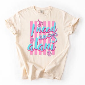 May include: An ivory short-sleeved t-shirt featuring a graphic print with vertical pink stripes. The text reads "I need an alani" in bright blue script. The design includes small pink illustrations of a cherry, a bow, and two strawberries.