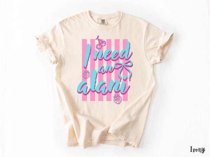May include: An ivory short-sleeved t-shirt featuring a graphic print with vertical pink stripes. The text reads "I need an alani" in bright blue script. The design includes small pink illustrations of a cherry, a bow, and two strawberries.