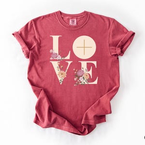 May include: Crimson t-shirt featuring the word "LOVE" in a cream font. The "O" is a circle with a cross, and the "V" and "E" are decorated with floral arrangements in pink, peach, and purple. The word "Crimson" is in the bottom right corner.