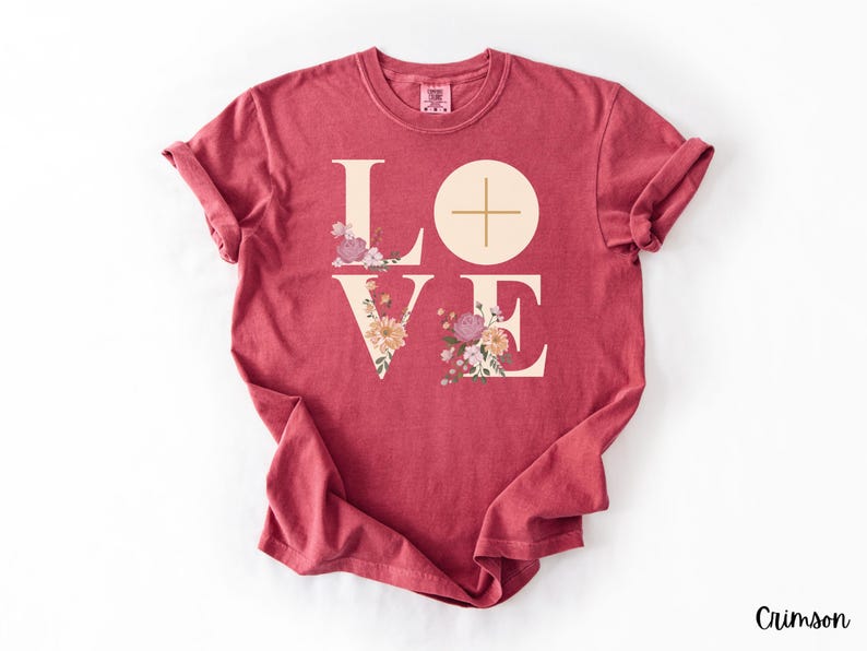 May include: Crimson t-shirt featuring the word "LOVE" in a cream font. The "O" is a circle with a cross, and the "V" and "E" are decorated with floral arrangements in pink, peach, and purple. The word "Crimson" is in the bottom right corner.