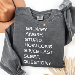 May include: A charcoal gray crewneck sweatshirt featuring white text that reads: GRUMPY. ANGRY. STUPID. HOW LONG SINCE LAST SLEEP, QUESTION? The garment has a relaxed fit with long sleeves and a ribbed neckline, showcasing a faded, vintage-inspired fabric finish.