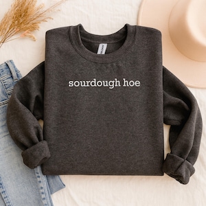 Peut inclure: Sweat-shirt gris foncé avec l'inscription blanche "sourdough hoe". Les manches et l'ourlet sont retroussés. Un chapeau clair et un jean bleu sont visibles en arrière-plan, sur une surface blanche.