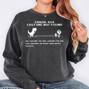 Puede incluir: Una sudadera gris carbón de cuello redondo con un gráfico pixelado blanco de un dinosaurio y un cactus. El texto dice: ERROR 404 COSTUME NOT FOUND. THE COSTUME YOU ARE LOOKING FOR WAS LOST, REMOVED OR MIGHT HAVE NEVER EXISTED.