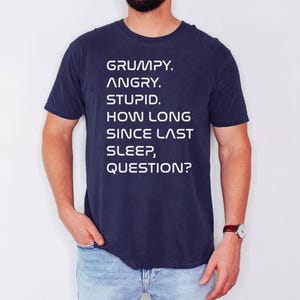 May include: Navy blue t-shirt with white text that reads: "GRUMPY. ANGRY. STUPID. HOW LONG SINCE LAST SLEEP, QUESTION?" The shirt is a short-sleeved crew neck.