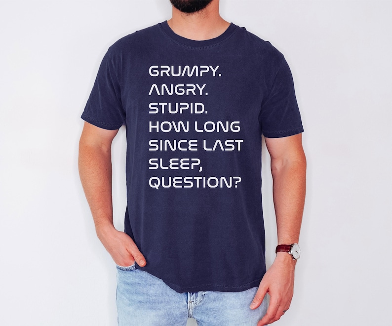 May include: Navy blue t-shirt with white text that reads: "GRUMPY. ANGRY. STUPID. HOW LONG SINCE LAST SLEEP, QUESTION?" The shirt is a short-sleeved crew neck.