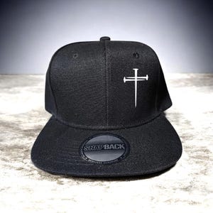 May include: Black snapback hat with a white cross design on the front. The hat has a flat brim and a snap closure at the back. The hat has the text "SNAPBACK" on the brim.