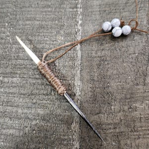 May include: A small, sharp, metal needle wrapped in brown cord with white beads on a brown cord attached.