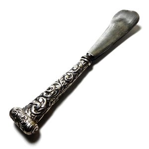 May include: A silver butter knife with an ornate handle. The handle features detailed scrollwork and a rounded end. The blade is a flat, rounded shape. The knife is set against a white background.