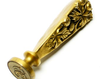 May include: A gold-colored antique wax seal stamp. The handle is decorated with a detailed relief of leaves and berries. The circular stamp head features an ornate monogram. The seal is set against a white background.