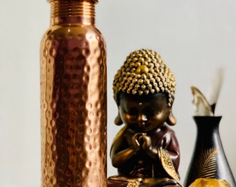 May include: A hammered copper water bottle with a traditional design sits next to a small golden statue of a meditating Buddha. A small yellow crystal and a black elephant figurine are also in the image.