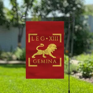 Könnte beinhalten: Eine rote Gartenflagge mit goldfarbenen Buchstaben und einem goldenen Löwenemblem. Der Text lautet "LEG XIII GEMINA" in einem rechteckigen Rahmen. Die Flagge hängt an einem schwarzen Metallständer.
