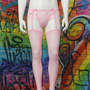 Può includere: Un manichino espone lingerie rosa, tra cui un reggicalze con fiocchi e calze trasparenti. Lo sfondo è un muro di mattoni dipinto con un vivace arcobaleno e graffiti.