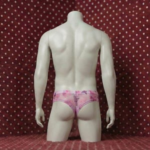 May include: A headless mannequin displays pink underwear with a floral pattern and butterfly accents. The underwear is sheer and has a low-rise cut. The mannequin is set against a red background with white polka dots.