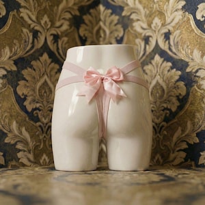 May include: A white ceramic sculpture of a lower torso with a pink ribbon and bow detail. The sculpture is set against a patterned background with gold, blue, and cream tones.