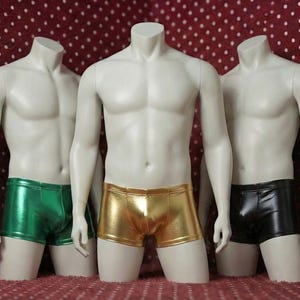 May include: Three mannequins wearing metallic shorts in green, gold, and black. The mannequins are headless and have a muscular physique. The background is a red fabric with white polka dots. The shorts are shiny and form-fitting.