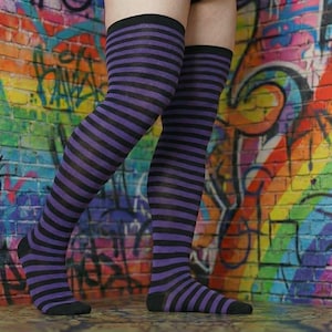 Pode incluir: Meias altas com listras pretas e roxas alternadas. As meias são usadas em um fundo de uma parede de tijolos coberta com grafites coloridos.