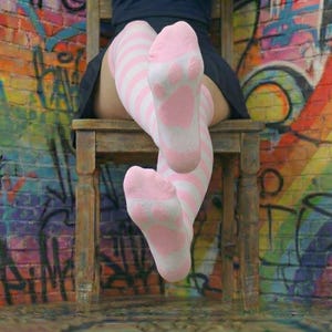 Könnte beinhalten: Kniehohe Socken mit rosa und weißen Streifen und Pfotenabdruckdetails. Die Socken werden beim Sitzen auf einem Holzstuhl getragen. Der Hintergrund ist eine Backsteinmauer mit farbenfrohen Graffiti.