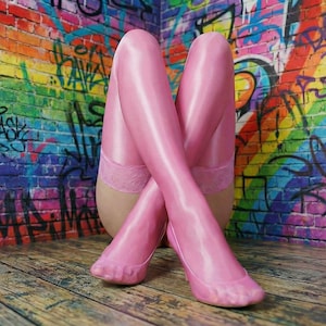 Può includere: Calze autoreggenti rosa con bordi in pizzo, gambe incrociate, contro un muro di mattoni coperto di graffiti. Il muro presenta un vibrante arcobaleno di colori e vari tag di graffiti. Il pavimento è fatto di assi di legno.