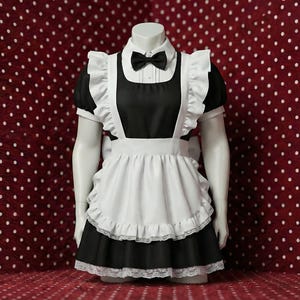 May include: A black and white maid costume displayed on a mannequin. The costume features a black dress with a white apron, ruffled trim, and a matching bow tie. The background is a red fabric with white polka dots.