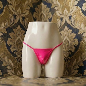 May include: A white ceramic form with a glossy finish, shaped to resemble a torso, is adorned with a vibrant pink thong. The background features a patterned wallpaper in shades of gold, blue, and brown.