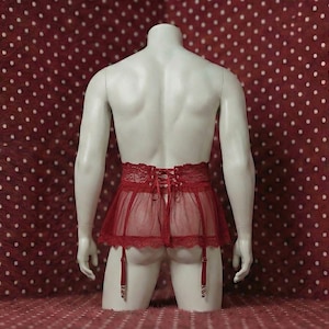 Può includere: Una gonna di lingerie rossa con finiture in pizzo e giarrettiere è esposta su un manichino. La gonna ha la schiena allacciata ed è realizzata in un tessuto trasparente. Il manichino è su uno sfondo rosso a pois bianchi.