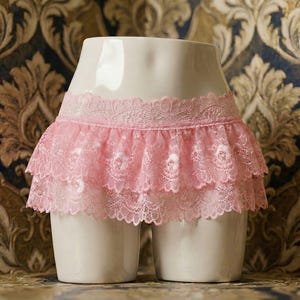 May include: A white mannequin torso with a pink lace skirt. The skirt has two layers of ruffled lace with a floral pattern. The background is a patterned wallpaper with gold and blue tones.